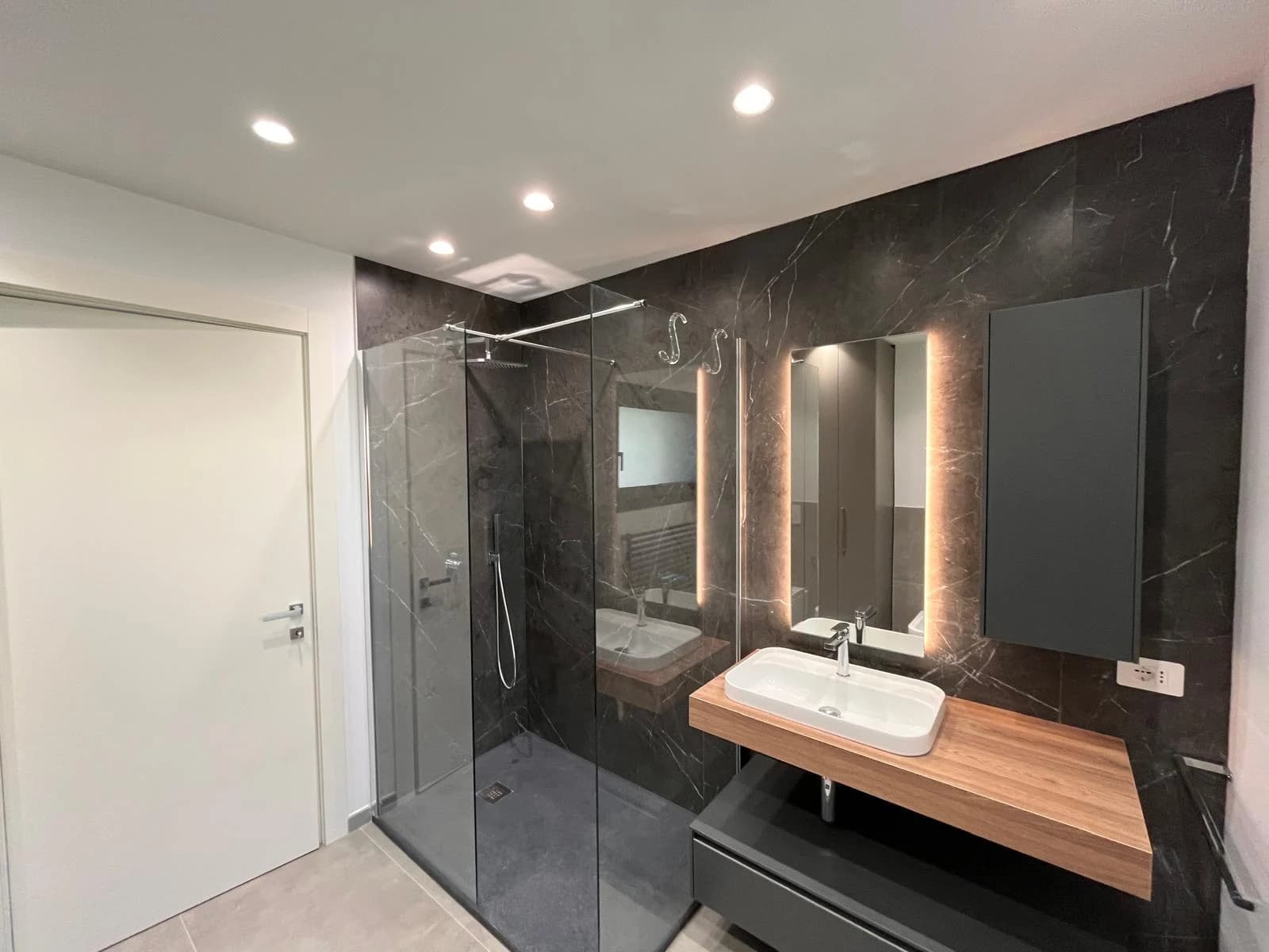 Il bagno con rivestimento scuro, doccia in vetro e specchio retroilluminato